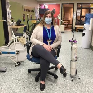 Masked woman sitting on chair with legs crossed surrounded by exercise equipment and oxygen tank